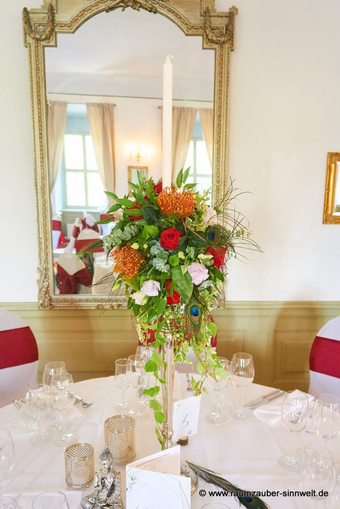 Tischdekoration aus roten Rosen, Nadelkissen, Eustoma, Hypericum und Pfauenfedern.