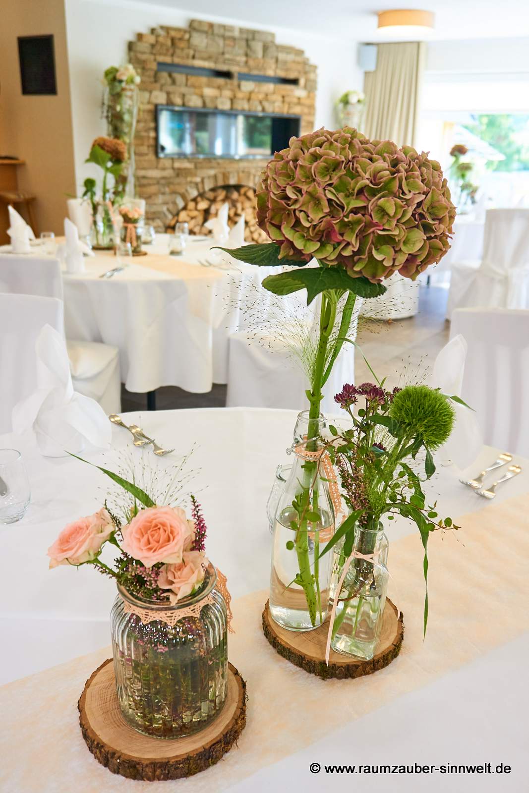 Tischdekoration aus Hortensie, Astrantie, Bartnelke, Rosen und Gräsern in Gläsern auf Baumscheibe