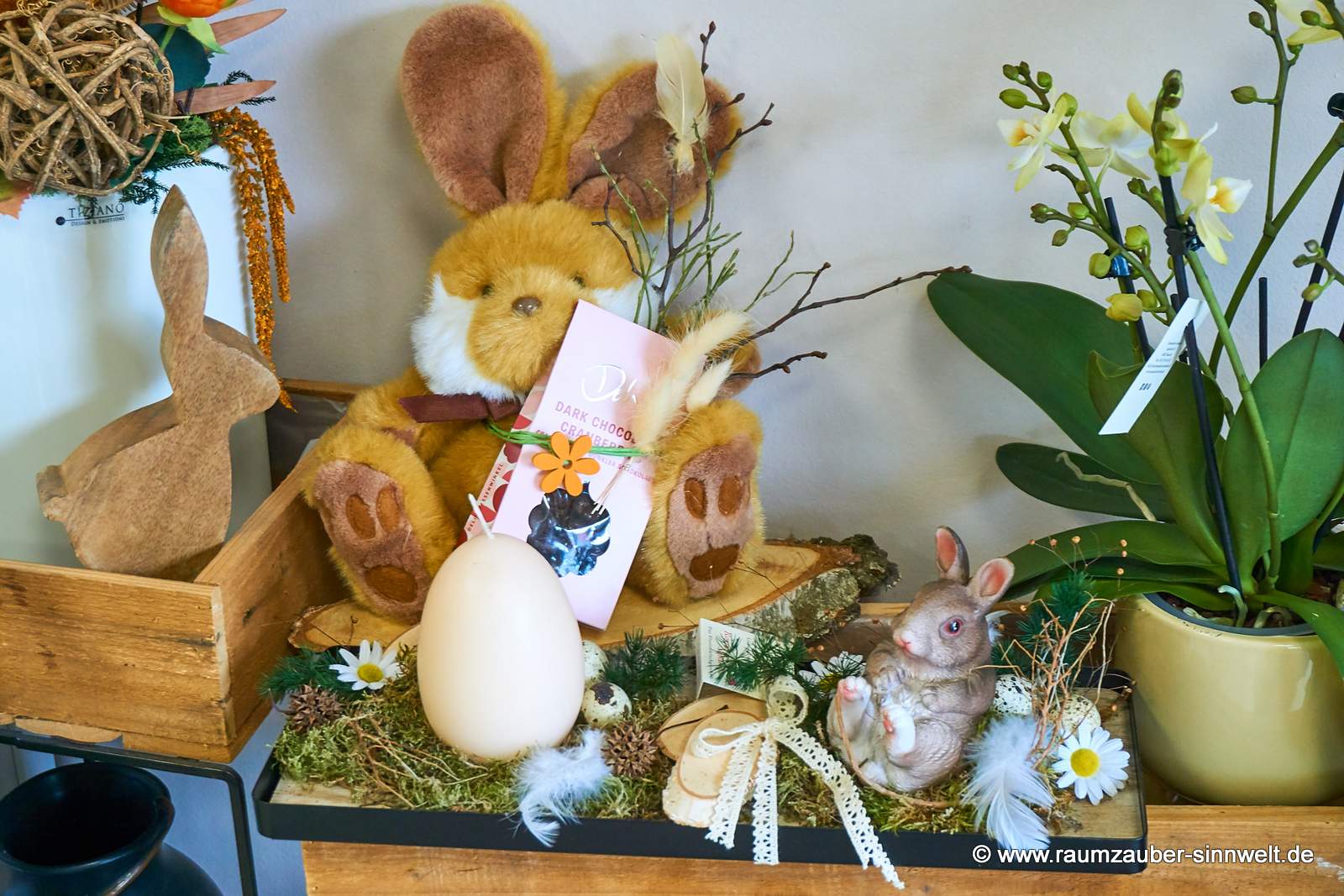 Osterdekoration mit Kerze und Dekohase, dahinter Präsent aus Stoff-Hasen mit Schokofrüchten dekoriert.